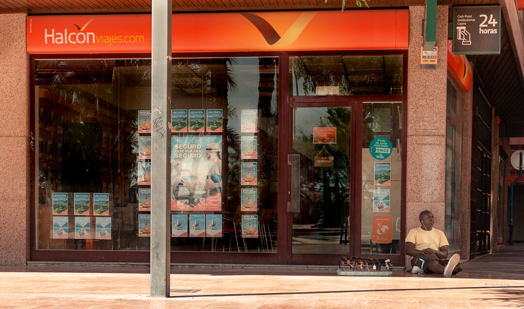 A man sitting on the pavement beneath a travel agency storefront in Alicante